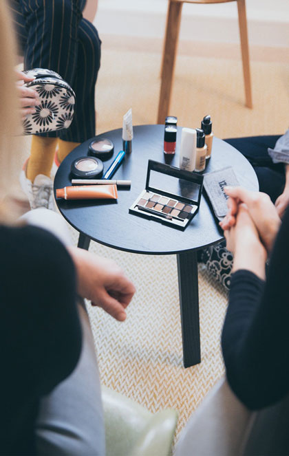 Woman looking in a brightly lit mirror, applying mascara to her upper lashes. A small, simple collection of beginner makeup products is neatly arranged on the vanity beside her.